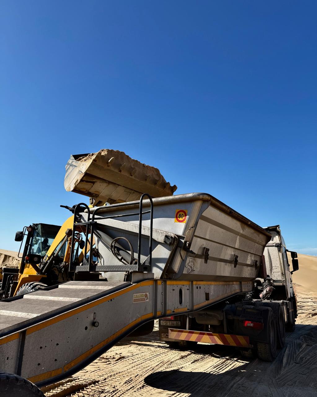 Komatsu front loader tipping sand into CWG truck against vivid blue sky